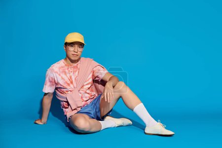 A trendy young man, stylishly dressed, sits on the ground wearing a vibrant yellow hat against a blue backdrop.