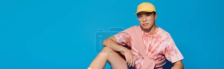 A stylish, young man sits on floor against a blue backdrop, sporting a yellow hat and trendy attire.