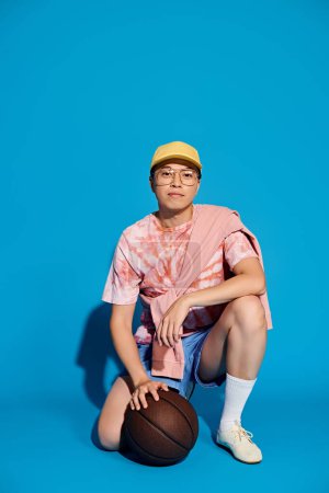 Ein stylischer junger Mann in modischer Kleidung sitzt auf dem Boden und hält selbstbewusst einen Basketball vor blauer Kulisse.