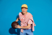 A fashionable young man energetically holds a basketball in his right hand against a blue backdrop. hoodie #699581050