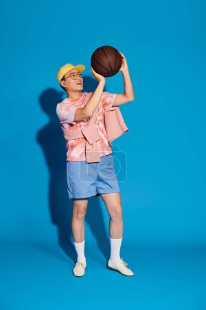 Stylish young man confidently holds a basketball in his right hand, exuding athleticism and coolness against a blue backdrop.