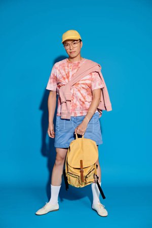 A trendy young man in a pink shirt and blue shorts poses with a yellow backpack against a vibrant blue backdrop.