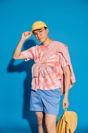 A stylish young man poses on a blue backdrop, wearing a pink shirt and blue shorts while holding a yellow bag.