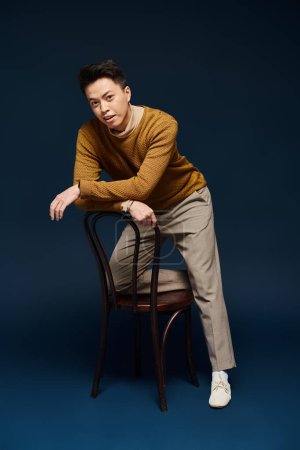 A fashionable young man in elegant attire confidently sits on top of a wooden chair, striking a dynamic pose.