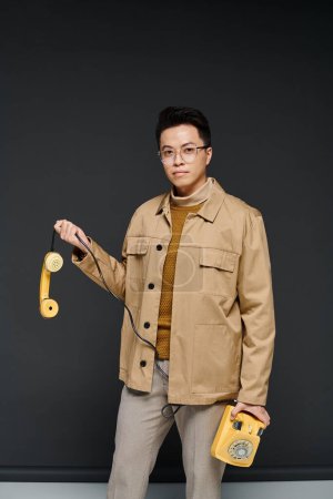 A fashionable young man poses actively while holding a yellow phone in his stylish tan jacket.