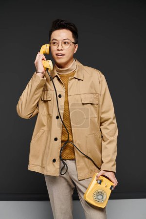 A fashionable young man in a tan jacket actively engages with a yellow phone.