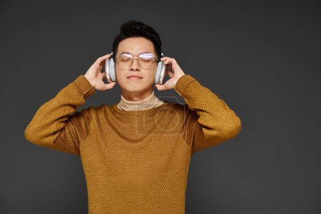 A stylish young man in elegant attire listening intently through headphones while wearing glasses.