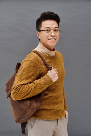 Foto de Un joven de moda con gafas y una mochila marrón posa con confianza en un atuendo elegante. - Imagen libre de derechos