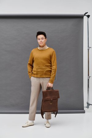 A fashionable young man stands confidently in front of a backdrop, holding a briefcase in a poised and assertive stance.