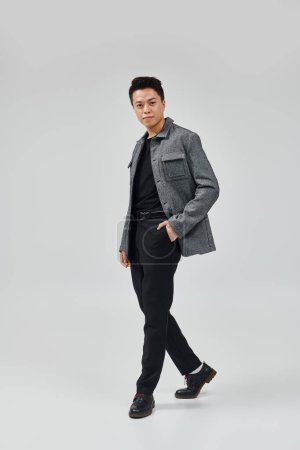 Téléchargez les photos : Un jeune homme à la mode pose activement dans une veste grise et un pantalon noir, exsudant élégance et style. - en image libre de droit