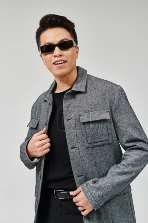 Ein modischer junger Mann posiert selbstbewusst in Mantel und Sonnenbrille und strahlt Eleganz und Raffinesse aus.