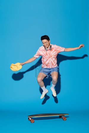 Un joven elegante salta con gracia en el aire mientras sostiene un monopatín, exudando energía y estilo sobre un telón de fondo azul.