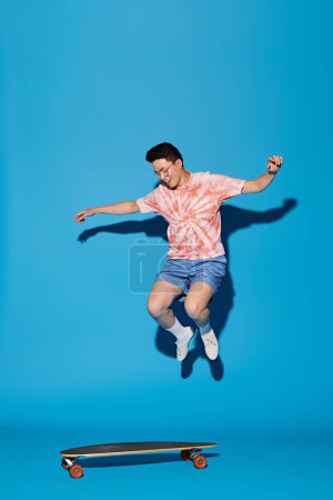 A stylish young man in trendy attire jumps in the air with a skateboard against a vibrant blue backdrop.