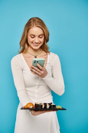 Una mujer rubia con gracia sostiene una bandeja de sushi en una mano y un teléfono celular en la otra, encarnando la elegancia multitarea.