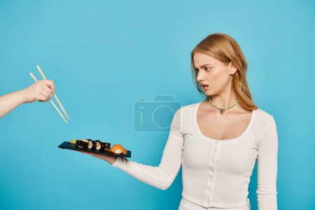 Photo for A beautiful blonde woman delicately holds a plate of sushi, showcasing the artistry of Japanese cuisine, chopsticks. - Royalty Free Image