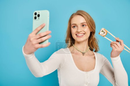 Une femme élégante avec des cheveux blonds tenant des sushis et des baguettes à la main et un téléphone portable dans l'autre.