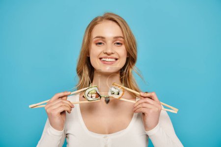Téléchargez les photos : Une femme blonde tient délicatement deux baguettes avec des sushis perchés dessus, mettant en valeur l'art élégant de savourer la cuisine japonaise. - en image libre de droit