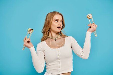 A woman with blonde hair gracefully holds two chopsticks with sushi perched on them.