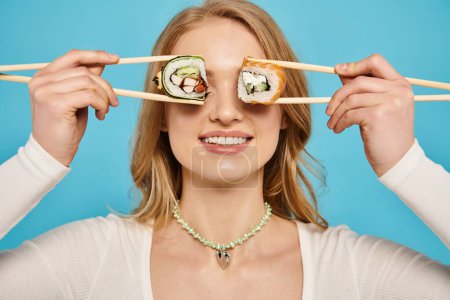 Blonde Frau hält spielerisch Essstäbchen über ihre Augen, auf denen Sushi thront, was einen lustigen und fantasievollen Moment darstellt.