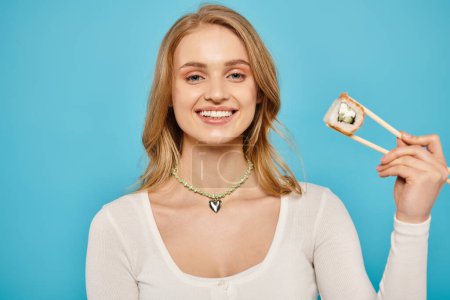 A beautiful woman with blonde hair holds a chopsticks with sushi in her right hand