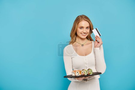 Photo for A beautiful blonde woman showcasing a tray filled with delicious sushi pieces, holding credit card. - Royalty Free Image