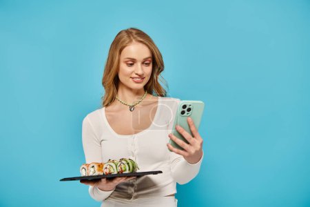 Una mujer elegante con el pelo rubio sosteniendo un plato de sushi y un teléfono celular, golpeando una pose.