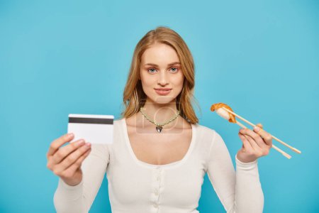 Una mujer rubia sostiene con confianza una tarjeta de crédito mientras muestra un delicioso sushi asiático.