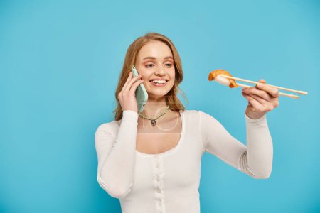 Eine blonde Frau hält zart Essstäbchen in der Hand, während sie am Telefon plaudert und asiatische Küche genießt.