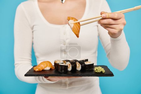 Cropped view of woman elegantly holds a plate of sushi and chopsticks, savoring each bite with a serene expression.