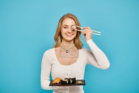 Eine schöne blonde Frau hält elegant einen Teller mit Sushi und Essstäbchen in der Hand und präsentiert die köstliche asiatische Küche.