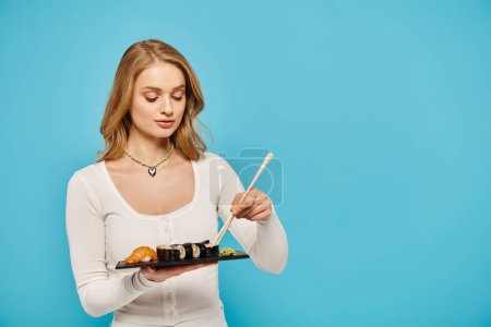A blonde woman gracefully holds a plate of sushi with chopsticks, showcasing the beauty of Asian cuisine.