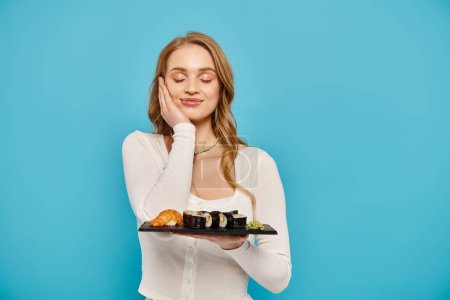 Une femme blonde tient gracieusement un plateau de sushi sur un fond bleu serein.