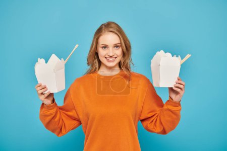 Téléchargez les photos : Une femme aux cheveux blonds tenant deux boîtes de nourriture asiatique dans ses mains, à la recherche coûteuse et contemplative. - en image libre de droit