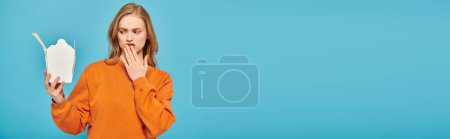 Téléchargez les photos : Une belle femme aux cheveux blonds tient une boîte de nourriture asiatique tout en portant une chemise orange. - en image libre de droit