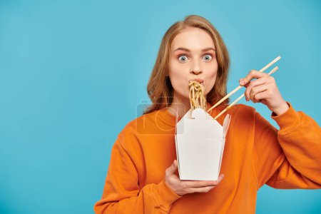 A beautiful woman with blonde hair delicately holds chopsticks in front of her mouth, savoring delicious Asian cuisine.
