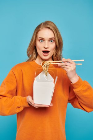 Une femme blonde chic tient élégamment des baguettes et une boîte de nouilles, mettant en valeur une appréciation pour la cuisine asiatique.