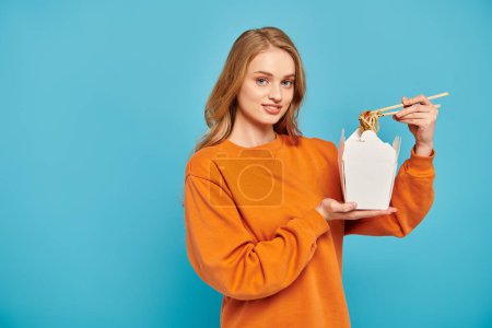 A stylish woman in an orange sweater holds box of Asian food.