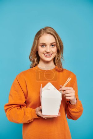 Eine blonde Frau im orangefarbenen Pullover wiegt eine Schachtel mit asiatischer Küche und strahlt Ruhe und kulinarische Wertschätzung aus..