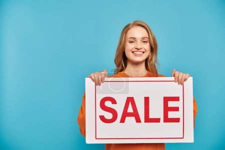 A blonde woman confidently holds a sign that reads Sale against a soft background.