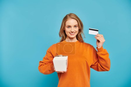 Una mujer elegante aparece contenta mientras sostiene una tarjeta de crédito en una mano y una caja de comida en la otra, simbolizando las compras en línea.