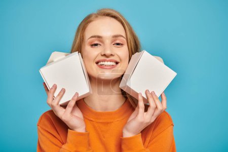 Una hermosa mujer rubia sostiene dos cajas de comida frente a su cara, ocultando su expresión con intriga.
