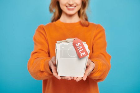 Vista recortada de mujer elegante sosteniendo caja de comida con una etiqueta de venta, mirando encantado e intrigado por el contenido.