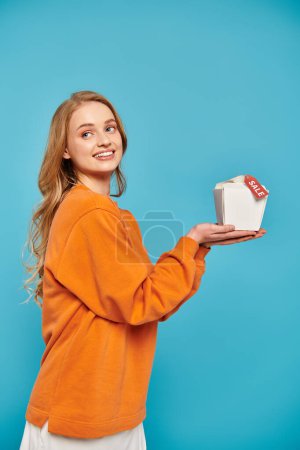 Eine elegante blonde Frau hält anmutig eine Lebensmittelschachtel mit Verkaufsschild in der Hand und strahlt Eleganz und Raffinesse aus.
