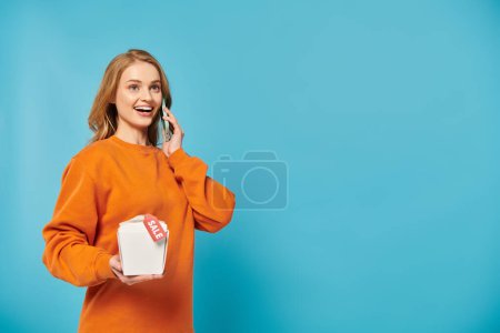 Una mujer con estilo en un suéter naranja se involucra en una animada conversación en un teléfono celular con caja de comida en la mano.