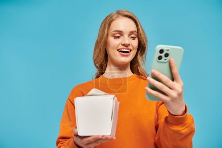 A blonde woman stylishly poses balancing a box of Asian food and a cell phone in her hands.