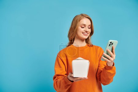 Une femme blonde équilibre sans effort une boîte de nourriture asiatique dans une main tout en faisant défiler son téléphone portable avec l'autre.