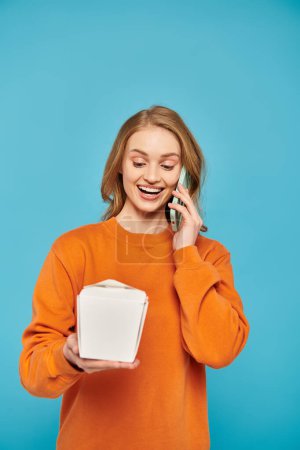 Photo for A stylish woman with blonde hair holds a box of Asian food while chatting on her cell phone. - Royalty Free Image