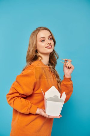 A stylish woman in an orange shirt holds food box and chopsticks