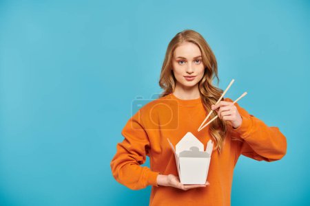 Eine schöne blonde Frau hält zart Essstäbchen und eine Schachtel leckeres asiatisches Essen in der Hand.