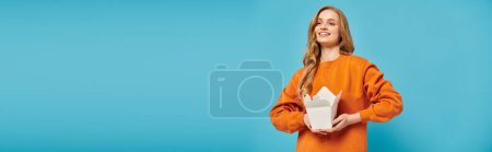 Téléchargez les photos : Une femme blonde dans un sweat-shirt orange tenant une boîte de nourriture avec un sourire sur le visage. - en image libre de droit